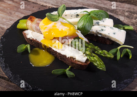 Toast con bastoni di asparagi verdi grigliati avvolto con pancetta, Uovo affogato , germogli e formaggio blu su nero ardesia pietra su vecchio rustico in legno Foto Stock