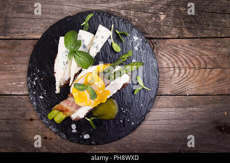 Toast con bastoni di asparagi verdi grigliati avvolto con pancetta, Uovo affogato , germogli e formaggio blu su nero ardesia pietra su vecchio rustico in legno Foto Stock