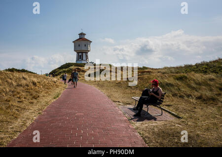 Water Tower, Langeoog. Germania, Deutschland. Un sentiero conduce verso una attrazione turistica, l'iconica white water tower - Wasserturm. Una giovane donna con i capelli rossi, si siede su un banco di pubblico per controllare il suo telefono cellulare. L'erba sulle dune di sabbia sono stati sbiancati dal sole durante l'estate di onda di calore. Foto Stock