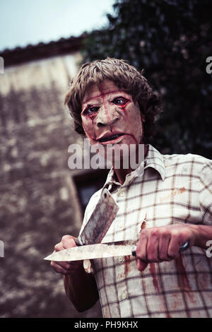 Un pauroso sfigurato uomo affilatura di un arrugginito e sanguinosa cleaver con un arrugginito e coltello insanguinato, di fronte a una casa in rovina in un inquietante paesaggio Foto Stock