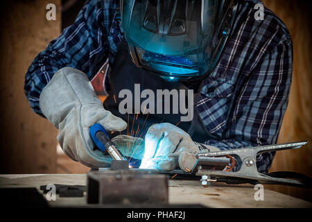 Close up di un giovane saldatore in uniforme, la maschera di saldatura e i saldatori pelli, metallo di saldatura con una macchina saldatrice ad arco in officina, blu e arancione Foto Stock