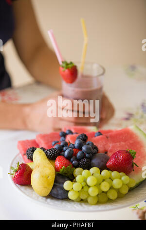 Close up estate deliziosa frutta organica sulla piastra con una donna su sfondo. Donna che mantiene un berry smoothie nelle mani e set di frutta per d Foto Stock