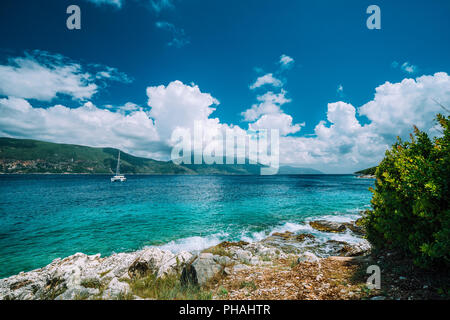 Crystal clear trasparente blu turchese teal mare mediterraneo acqua in Fiskardo città. White yacht in mare aperto al di ancoraggio sotto Incredibili nuvole bianche, CEFALLONIA, ISOLE IONIE, Grecia Foto Stock