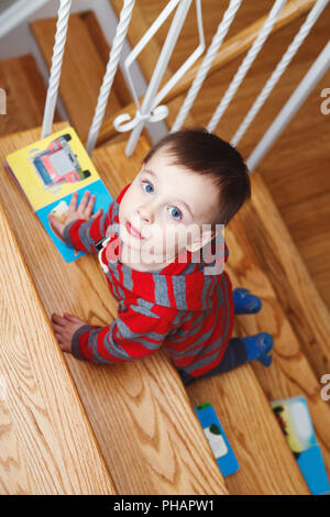 Ritratto od carino adorable little boy bimbo di due anni con libri per bambini arrampicate sulle scale per interni all'interno guardando in alto, overhead shot superiore Foto Stock