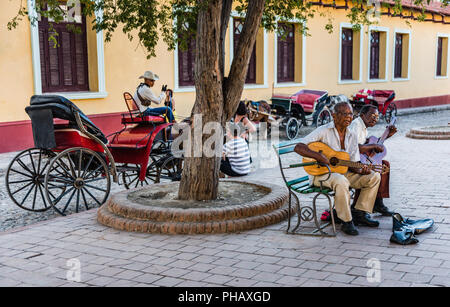 Trinidad, Cuba / Marzo 15, 2016: due chitarristi cubano seduta su una panchina la riproduzione di chitarre in una piazza nell'UNESCO città di Trinidad; una fila di cavallo e bu Foto Stock