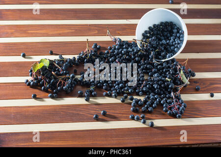 Ciotola piena di aronia versato sul tavolo di legno Foto Stock