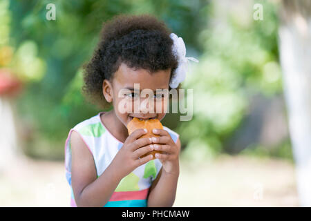 Una bella 4 anno vecchio African American Girl tenendo a due mani patty. Un piccolo di carnagione scura ragazza allegramente mangia il pane. Foto Stock