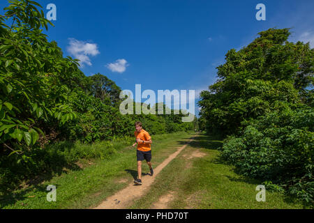 Un giovane cacuasiano in una tee arancione corre su un sentiero in un ambiente forestale. Singapore. Foto Stock