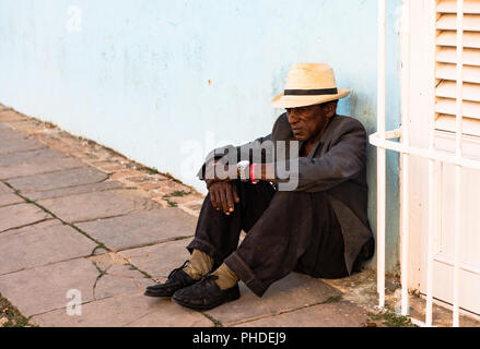 Trinidad / Cuba - Marzo 15, 2016: indigenti uomo cubano, indossando la paglia fedora e ben indossato tuta, siede sulla terra contro la parete blu in plaza square. Foto Stock