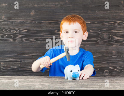 Un piccolo cinque-anno-vecchio ragazzo con i capelli rossi vuole rompere il suo salvadanaio con un martello per ottenere la sua moneta risparmio, primo piano Foto Stock