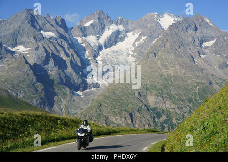 Motociclista in crociera con le alte cime e ghiacciai del Massiccio degli Ecrins sullo sfondo. Tra col du Lautaret e col du Galibier, Francia. Foto Stock