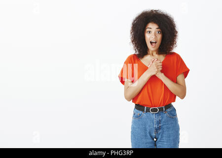 Piscina colpo di stupiti lieta donna attraente con la pelle scura e capelli ricci, stringendo le mani e fissando con soddisfazione le emozioni, essendo affascinato con interessanti foto su sfondo grigio Foto Stock