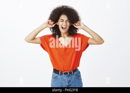 Studio shot della bella ragazza africana con taglio di capelli afro, tenendo le dita negli orecchi, chiudendo gli occhi e gridando mentre è sotto pressione, avente in lotta con il mio ragazzo e non vogliono sentire risiede Foto Stock