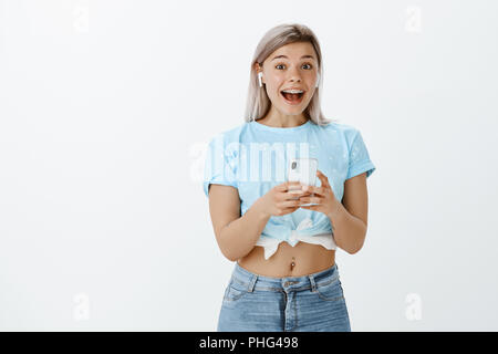 Affascinante emtoive giovane donna in abito elegante, tenendo smarpthone e ascoltando musica in auricolari wireless, sorridente largamente, essendo eccitato e gioioso audizione nuova via del cantante preferito Foto Stock