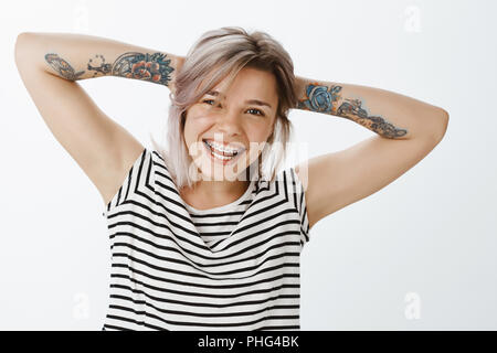 Essere felice e non mollare mai. Carefree attraente europea femminile con capelli biondi e tatuaggi, tenendo le mani dietro la testa, in piedi in posa rilassata e sorridente largamente, mostrando shiny carino bretelle Foto Stock