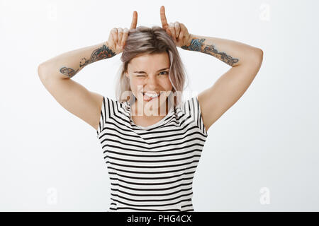 Testardo bella ragazza con capelli biondi e tatuaggi, winking e sorridente largamente, mostrando carino rinforzi su denti, rendendo le corna da indice delle dita sulla testa, essendo giocoso e flirty Foto Stock