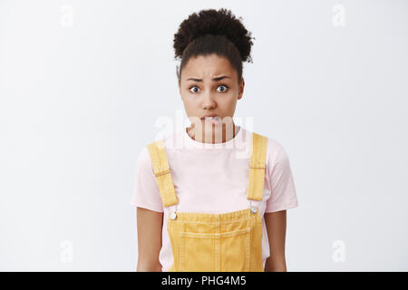 Oops, ho fatto errore, sentendosi in colpa. Ritratto di preoccupato insicuro afro-donna americana con bun hairstyle, mordere il labbro nervosamente e guardando la telecamera, che vogliono dire dispiace oltre il muro grigio Foto Stock