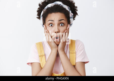 Close-up shot di sconvolti e entusiasta affascinante afro-americano di giovani collega di sesso femminile nella fascia oltre i capelli ricci e tuta gialla, soffocare, labbra di piegatura da stupore, guardando la telecamera impressionato Foto Stock