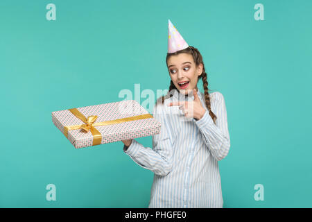 Sorpreso funny girl in striped maglietta blu e tappo di compleanno sulla testa piedi, tenendo cercando e che puntano a punteggiato confezione regalo con amazed faccia felice. Foto Stock