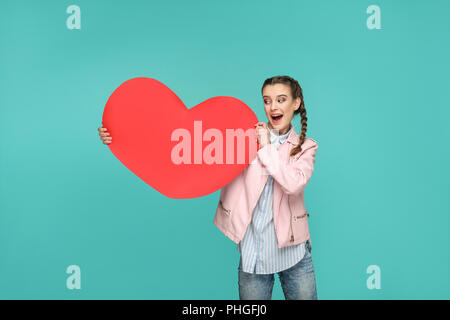 Stupito bella ragazza in stile casual, pigtail peli e giacca, permanente e la detenzione e guardando Big Red in forma di cuore con sorpresa di fronte. interni, Foto Stock