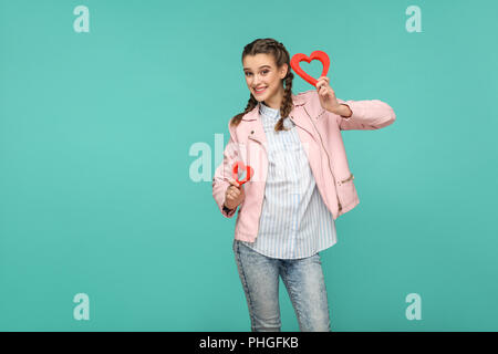 Felice bella ragazza in stile casual, pigtail i capelli e la camicia rosa, permanente e la holding cuore rosso forme e guardando la telecamera con un sorriso toothy Foto Stock