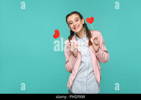 Felice bella ragazza in stile casual, pigtail pettinatura e camicia rosa, permanente e la holding cuore rosso adesivi e guardando la fotocamera e toothy smil Foto Stock