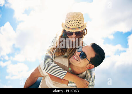 Bel giovane uomo con la sua amorevole moglie o fidanzata avendo divertimento rilassante per la vacanza estiva piggy back riding contro un soleggiato nuvoloso cielo blu Foto Stock