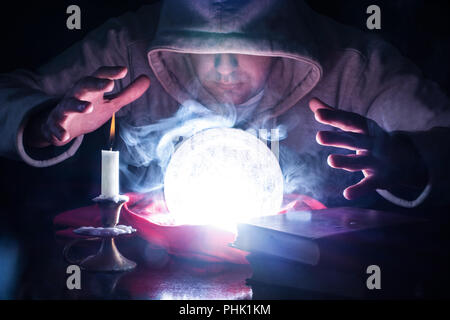 Procedura guidata con cappa e luci fumo magic sfera di cristallo sulla scrivania con candela in candeliere e vecchi libri predire il futuro guardando nella sfera Foto Stock