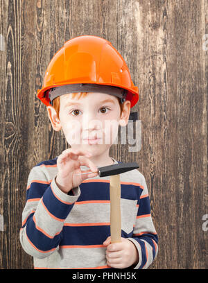 I capelli rossi boy in un casco arancione con un martello e un chiodo, primo piano su un nero lo sfondo di legno Foto Stock