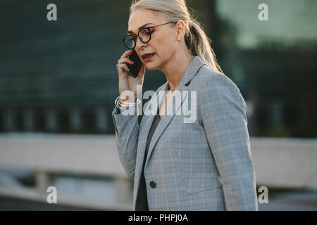 Ritratto di senior donna caucasica parlando al cellulare nella città. Femmina nel business suit passeggiate all'aperto parlando al telefono cellulare. Foto Stock