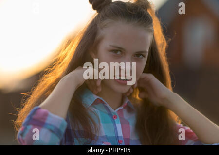 Felice ragazza adolescente guardando la fotocamera durante la regolazione marrone capelli. Sfocato edificio rosso in background. Foto Stock