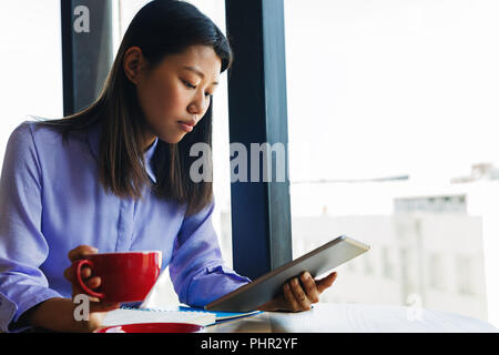 Vista laterale di una giovane donna asiatica tenendo una tazza di caffè e la lettura dalla sua tavoletta digitale Foto Stock