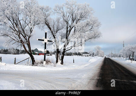 Paesaggio invernale che mostra una croce di legno da una strada e una fattoria con il pupazzo di neve alberi, bleu cielo, nuvole, paese recinto. Foto Stock