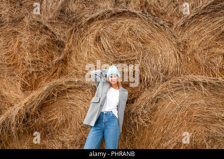 Piuttosto darkhaired fresco giovane donna camminare fuori nel sole contro lo sfondo di haystacks e indossa una tunica, maglia hat, jeans.Concetto di Foto Stock