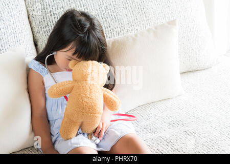 Carino bambina è la riproduzione medico con stetoscopio e orsacchiotto sensazione di stanchezza seduti sul divano di casa. Kid e health care concetto. Foto Stock