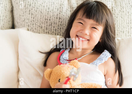 Carino bambina è sorridente e riproduzione medico con stetoscopio e orso di peluche. Kid e health care concetto. Foto Stock