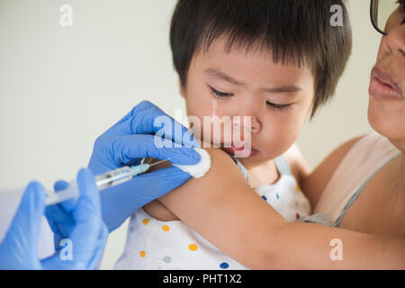 Medico dando un vaccino di iniezione ad una ragazza. Bambina piangendo con sua madre sullo sfondo. Foto Stock