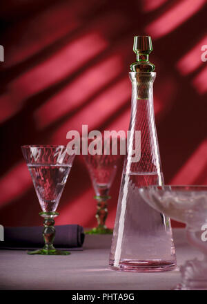 Decanter di vetro con acqua su un tavolo su uno sfondo rosso con raggi solari Foto Stock