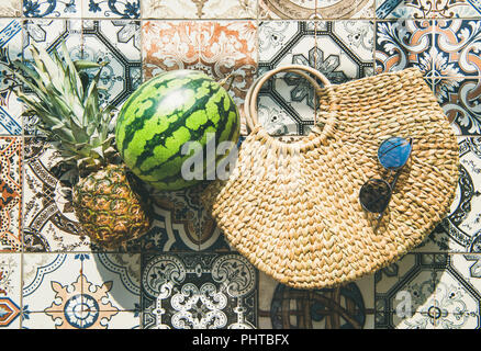 Estate di sfondo dello stile di vita. Flat-lay di frutta estiva di ananas e anguria, borsa di paglia e occhiali da sole su colorate piastrelle marocchine piano, vista dall'alto, Foto Stock