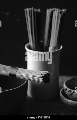 Soba Noodles, bambù vaporizzatore e cucina asiatica puntelli su sfondo scuro. Immagine in bianco e nero Foto Stock