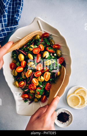 Una donna di agitazione di oliva verde con insalata di erbe e di mosti di uve i pomodori in una ciotola bianca con una foglia forma fotografato su sfondo grigio dalla vista dall'alto. Su Foto Stock