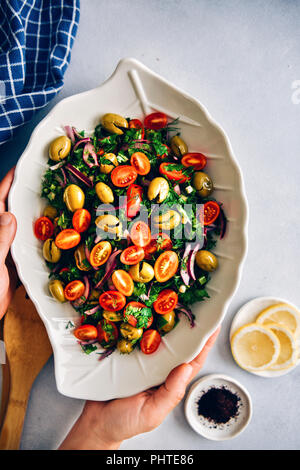 Una donna che serve verde verde oliva con insalata di erbe e di mosti di uve i pomodori in una ciotola bianca con una foglia forma fotografato su sfondo grigio dalla vista dall'alto. Somma Foto Stock