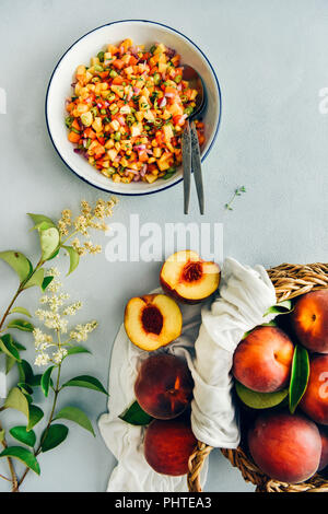 Peach salsa in una ciotola bianco, due cucchiai sul lato accompagnato da un cesto di pesche fresche e minuscoli fiori fotografati su sfondo grigio da t Foto Stock