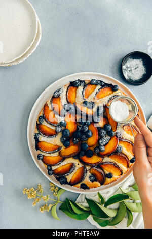 Una donna di vagliatura di zucchero a velo su una torta di pesche con mirtilli fotografato su sfondo grigio dalla vista dall'alto. Foglie verdi, un nero ciotola con powde Foto Stock