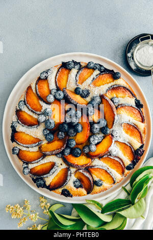 Torta di pesche con mirtilli spolverata con zucchero a velo fotografato su sfondo grigio dalla vista dall'alto. Foglie verdi e un setaccio con polvere Foto Stock