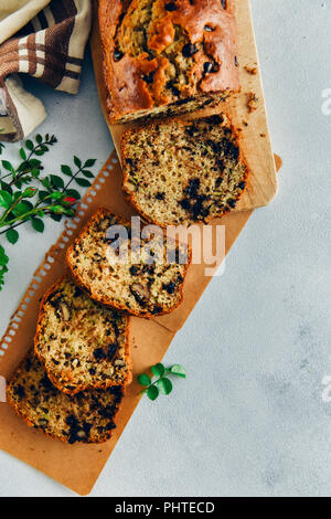 Con scaglie di cioccolato pane di zucchine con le noci tagliate a fette su un tagliere di legno e posta su carta marrone accompagnata da foglie verdi su sfondo grigio Foto Stock