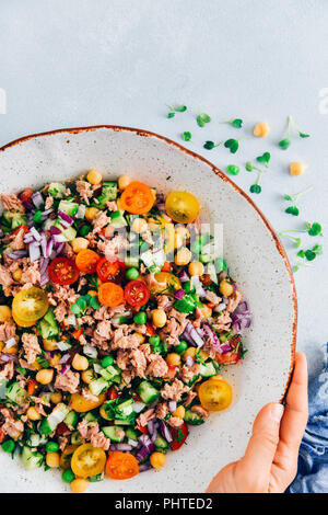 Mano che regge una ciotola di insalata di tonno con ceci, piselli, pomodori, cetrioli, cipolla e erbe aromatiche fotografata da vista dall'alto su sfondo grigio. Foto Stock