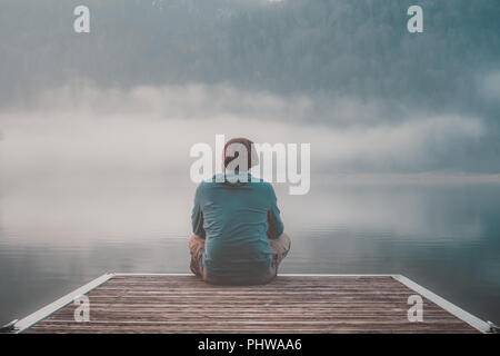 Giovane uomo con cappa di seduta sul molo di un lago in un giorno di nebbia. Foto Stock