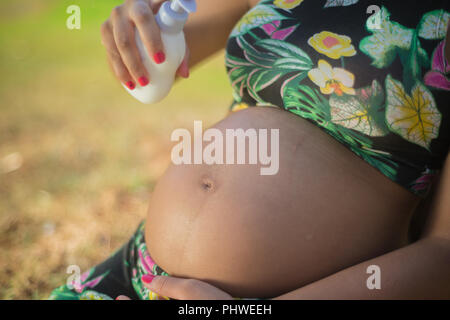 Donna incinta in un parco mettendo la crema sul suo ventre per evitare smagliature. Foto Stock