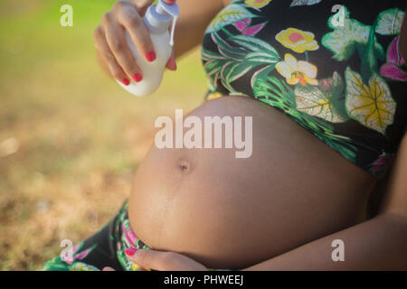 Donna incinta in un parco mettendo la crema sul suo ventre per evitare smagliature. Foto Stock